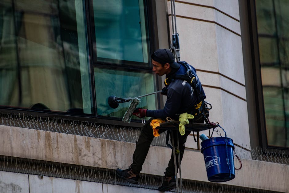 Man cleaning high-rise building windows using safety harness outdoors in daylight.